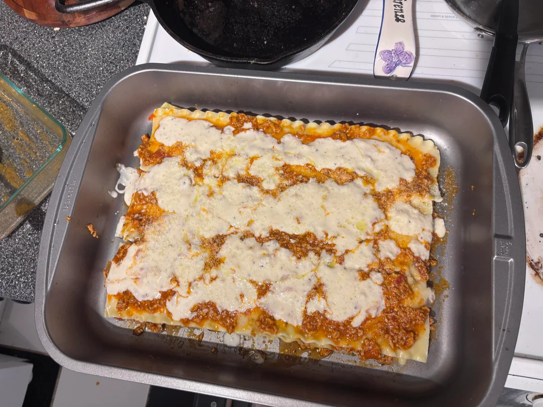 A rectangular pan of baked lasagna with layers of pasta, meat sauce, and melted cheese sits on a stovetop, next to a cast iron skillet and spatula.