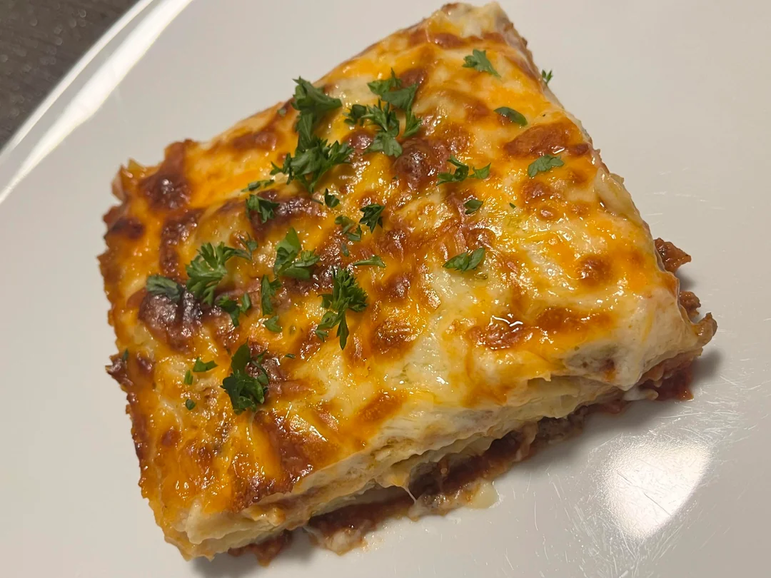 A square slice of lasagna with melted cheese and browned edges, topped with chopped parsley, served on a white plate.