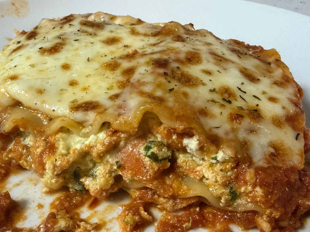 A close-up of a slice of lasagna with melted cheese on top, visible layers of pasta, tomato sauce, ricotta cheese, and herbs on a white plate.