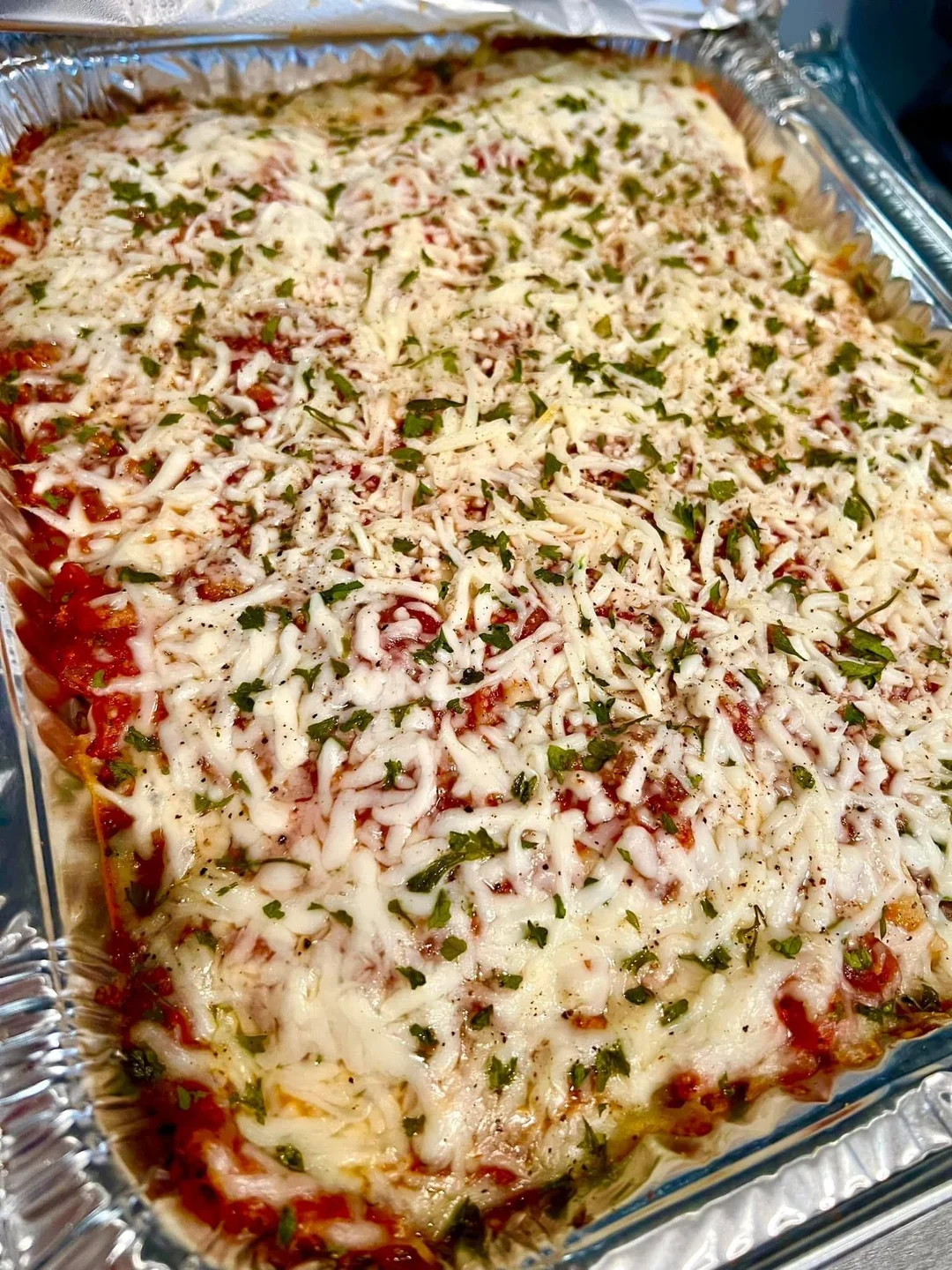 A large tray of baked lasagna topped with melted cheese, tomato sauce, and chopped herbs, displayed in an aluminum foil container.
