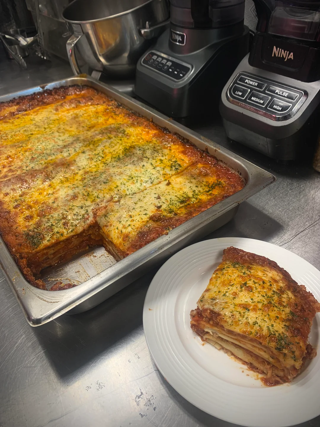 A baked lasagna in a metal pan with a slice removed and served on a white plate; kitchen appliances are visible in the background.