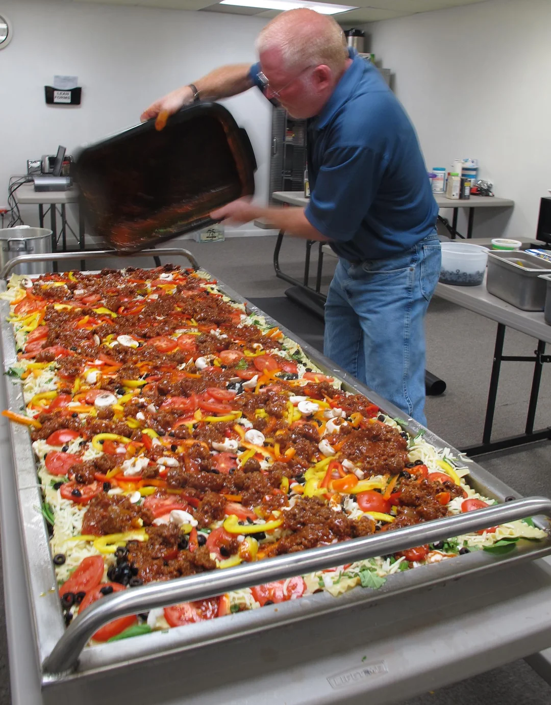 A man pours a pan of sauce onto a large rectangular pizza topped with vegetables and meat, in an industrial kitchen setting.