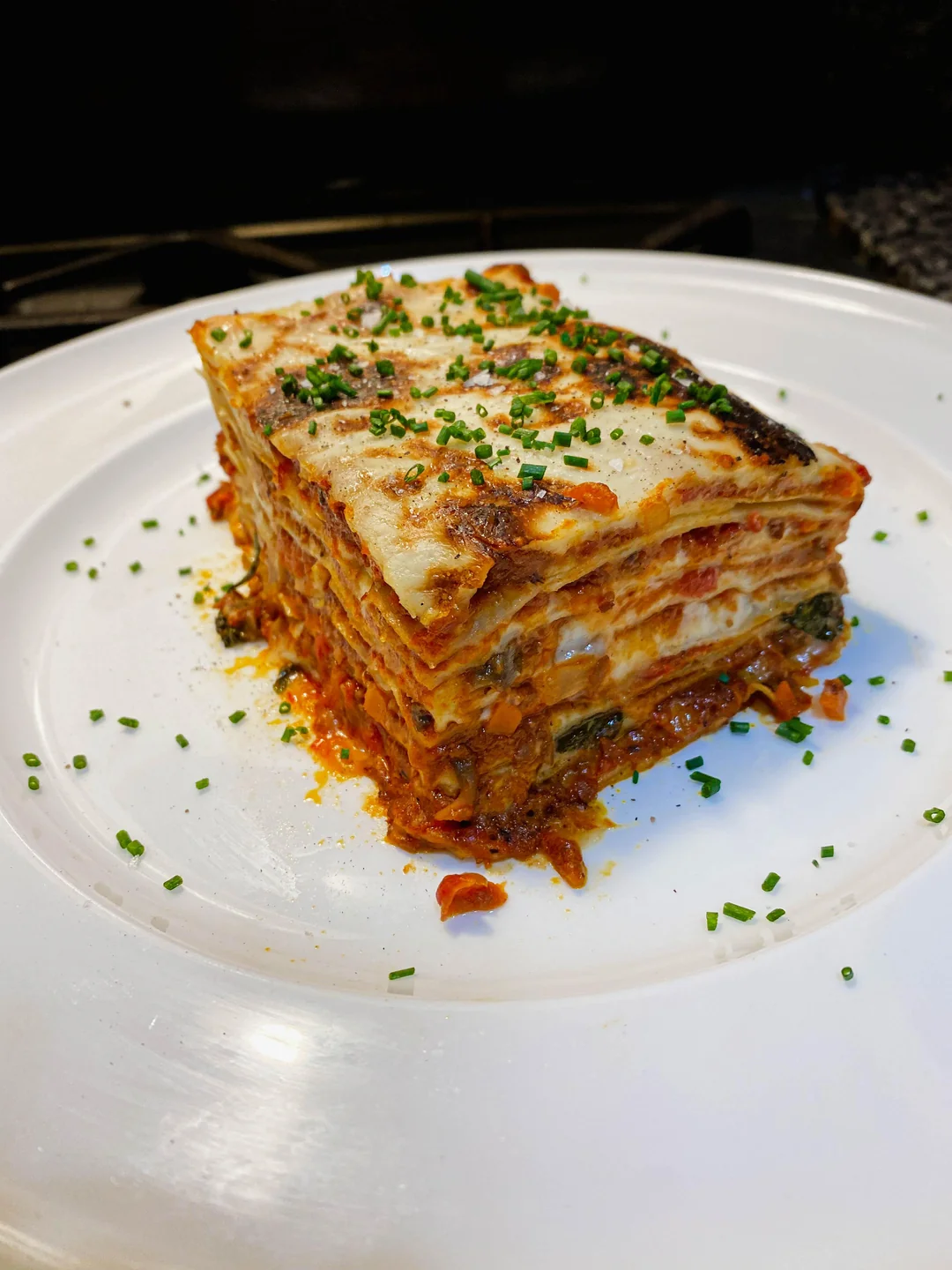 A thick, neatly cut slice of vegetable lasagna is served on a white plate, topped with chopped chives and visible layers of pasta, sauce, cheese, and vegetables.