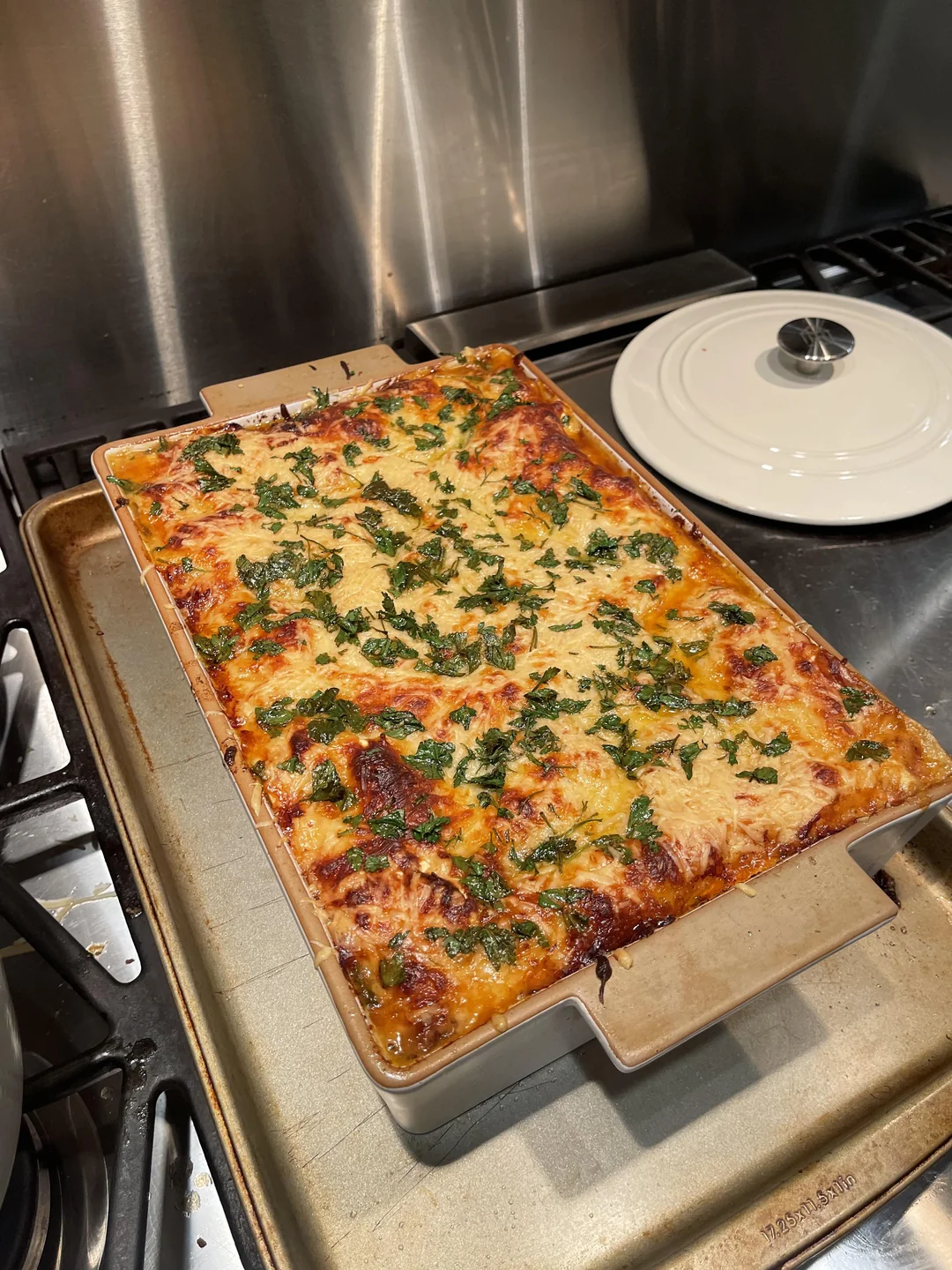 A freshly baked lasagna in a rectangular ceramic dish sits on a baking sheet atop a stove, with melted cheese and parsley visible on top. A white pot with a lid is in the background.
