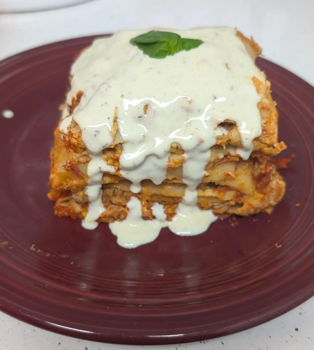 A slice of lasagna topped with creamy white sauce sits on a maroon plate, garnished with a single basil leaf.