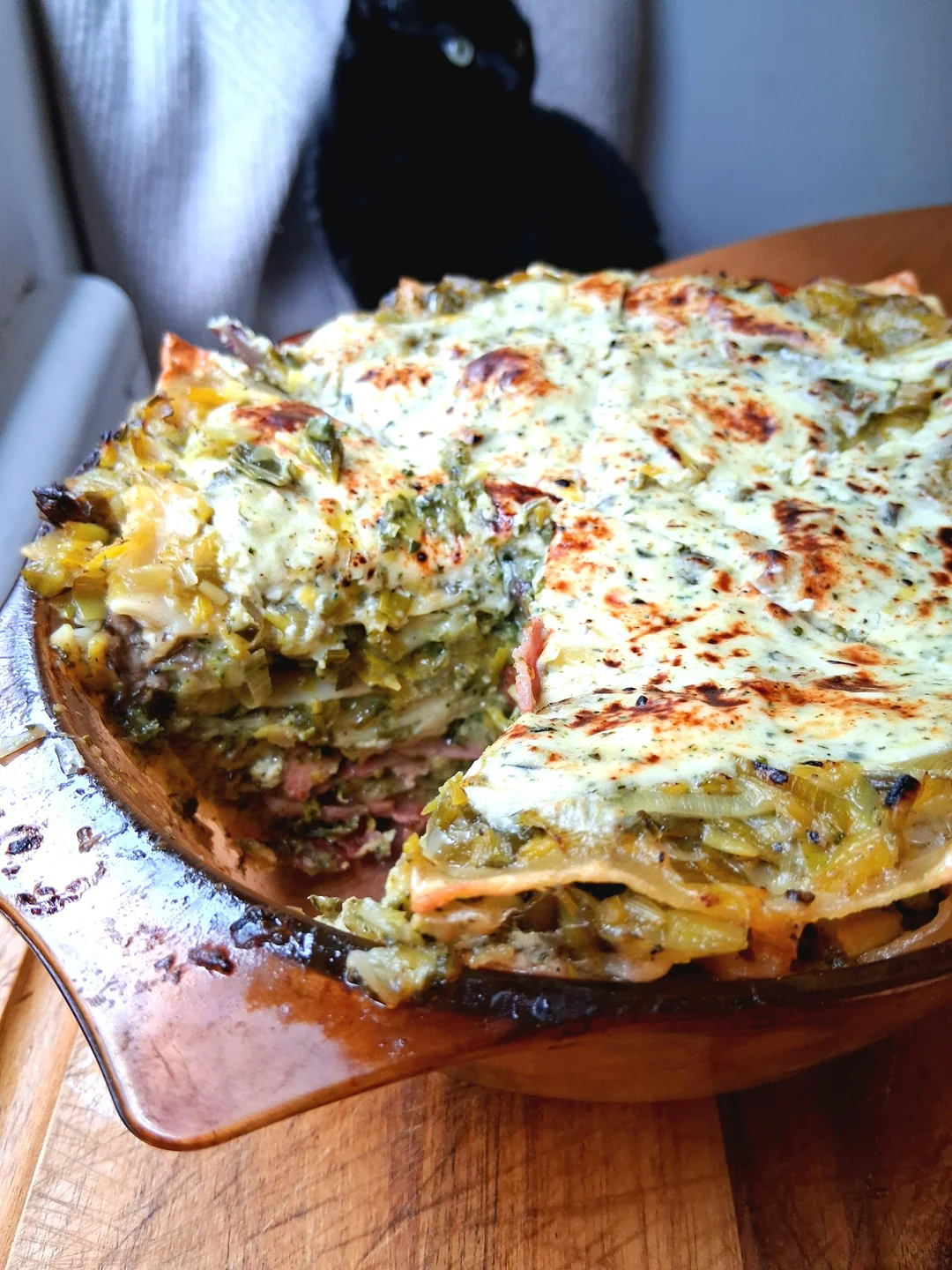 A baked lasagna with a slice removed, showing layered pasta, greens, sauce, and cheese. The dish sits on a wooden surface, with a black cat in the blurred background near a window.