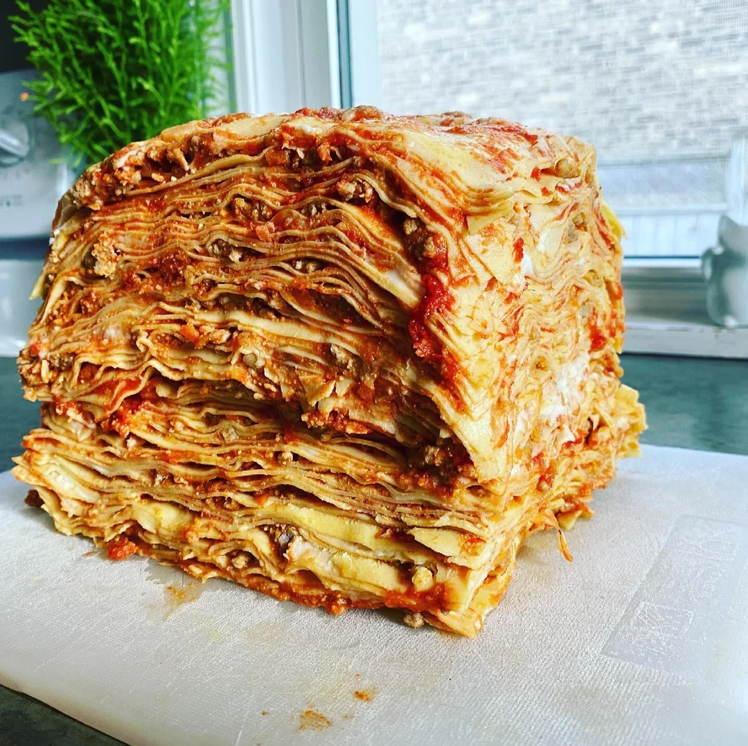 A large, thick, square stack of layered lasagna with visible sauce and cheese, sitting on a white cutting board by a window with a green plant in the background.