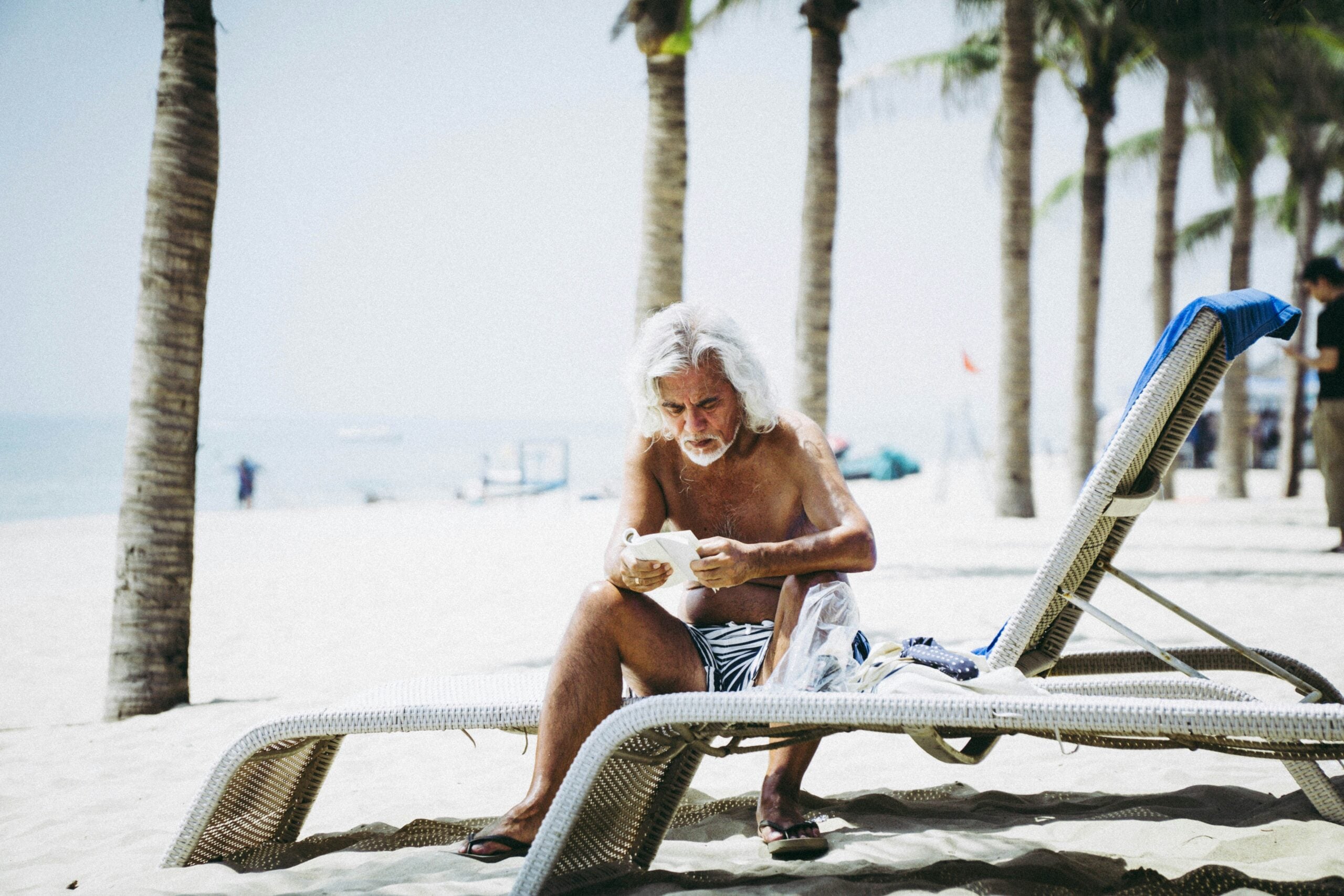 Older man with long white hair sits on a lounge chair under palm trees at a beach, reading a book or paper—perhaps exploring tips on boomers saving money. Sand, sea, and more beachgoers appear in the background on a sunny day.