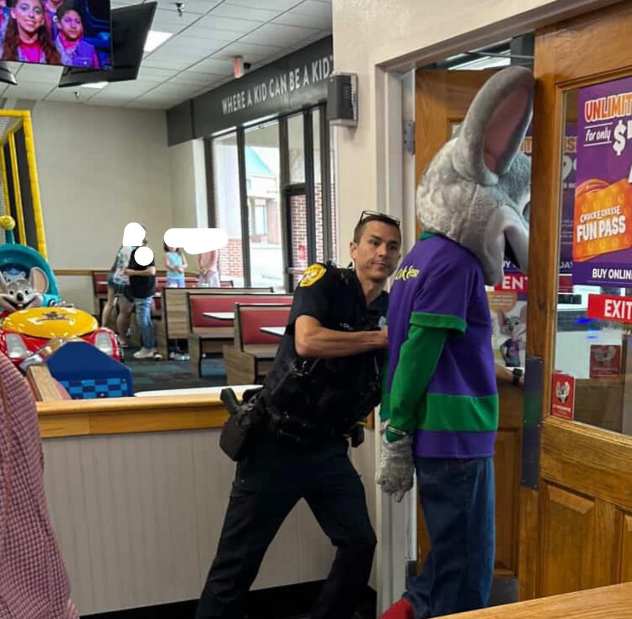 A police officer playfully pretends to arrest a person in a Chuck E. Cheese mascot costume near an exit door inside the restaurant.