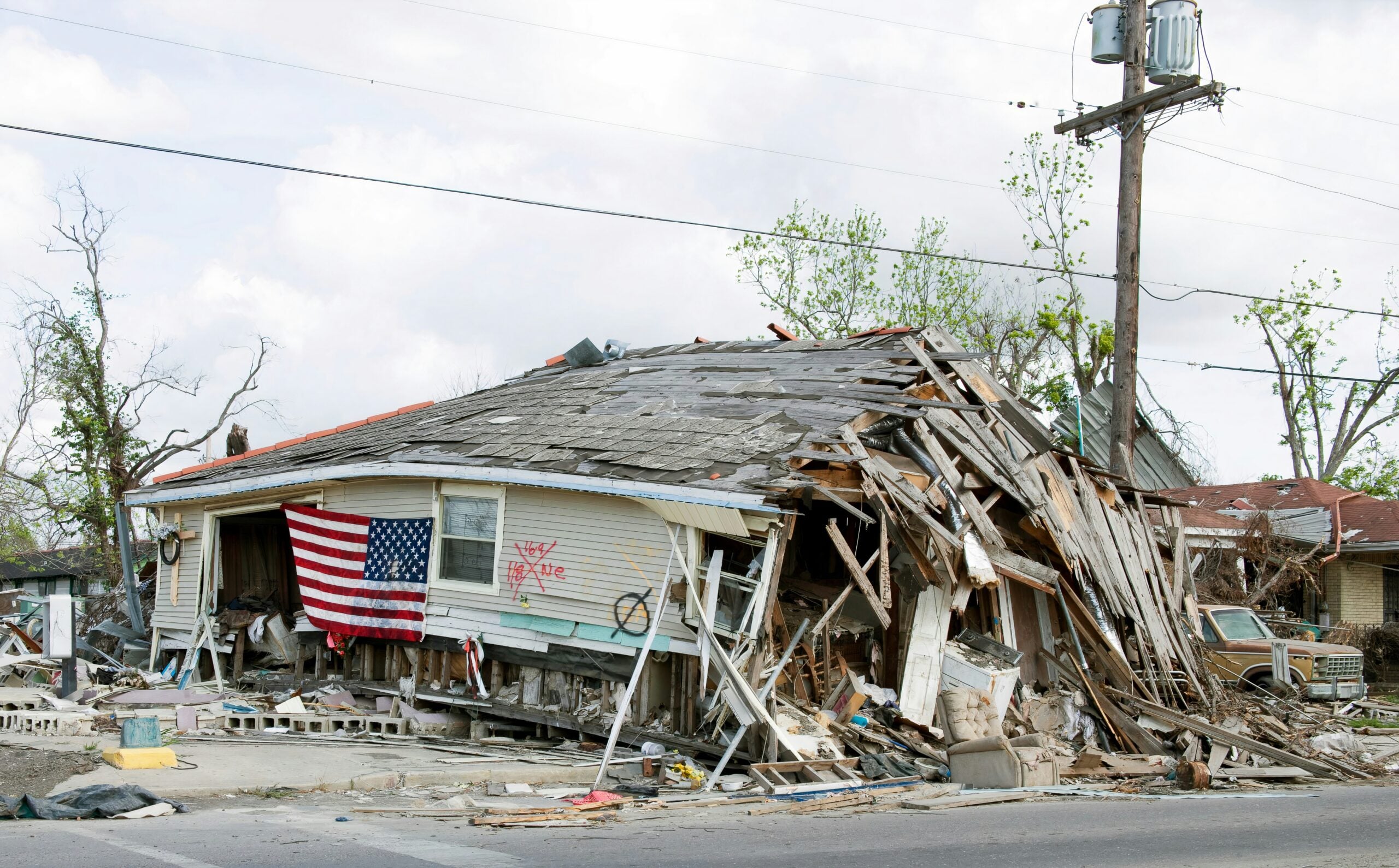 A house with severe structural damage, partially collapsed, displays an American flag on its porch. Debris and fallen trees surround the building, indicating recent destruction—possibly from a natural disaster—serving as a stark reminder of the challenges faced in life in 2026.