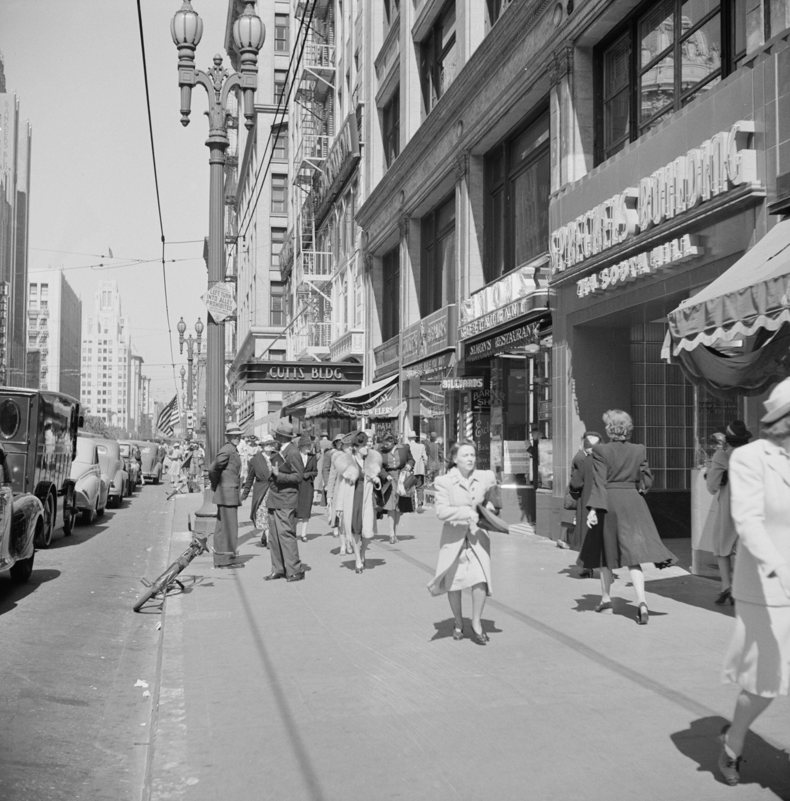 Black-and-white photo of a busy city sidewalk with people walking and shopping, parked cars, old-fashioned street lamps, and storefront signs. A girl in the foreground appears to be skipping or dancing.