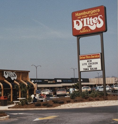 A vintage photo of a D’Lites fast food restaurant with a large sign advertising new lite chicken and tuna salad. Cars are parked in front and a shopping center is visible in the background.