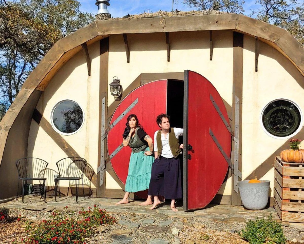 Two people dressed in fantasy costumes stand outside a hobbit-style house with a round red door, surrounded by plants and outdoor chairs, on a sunny day.