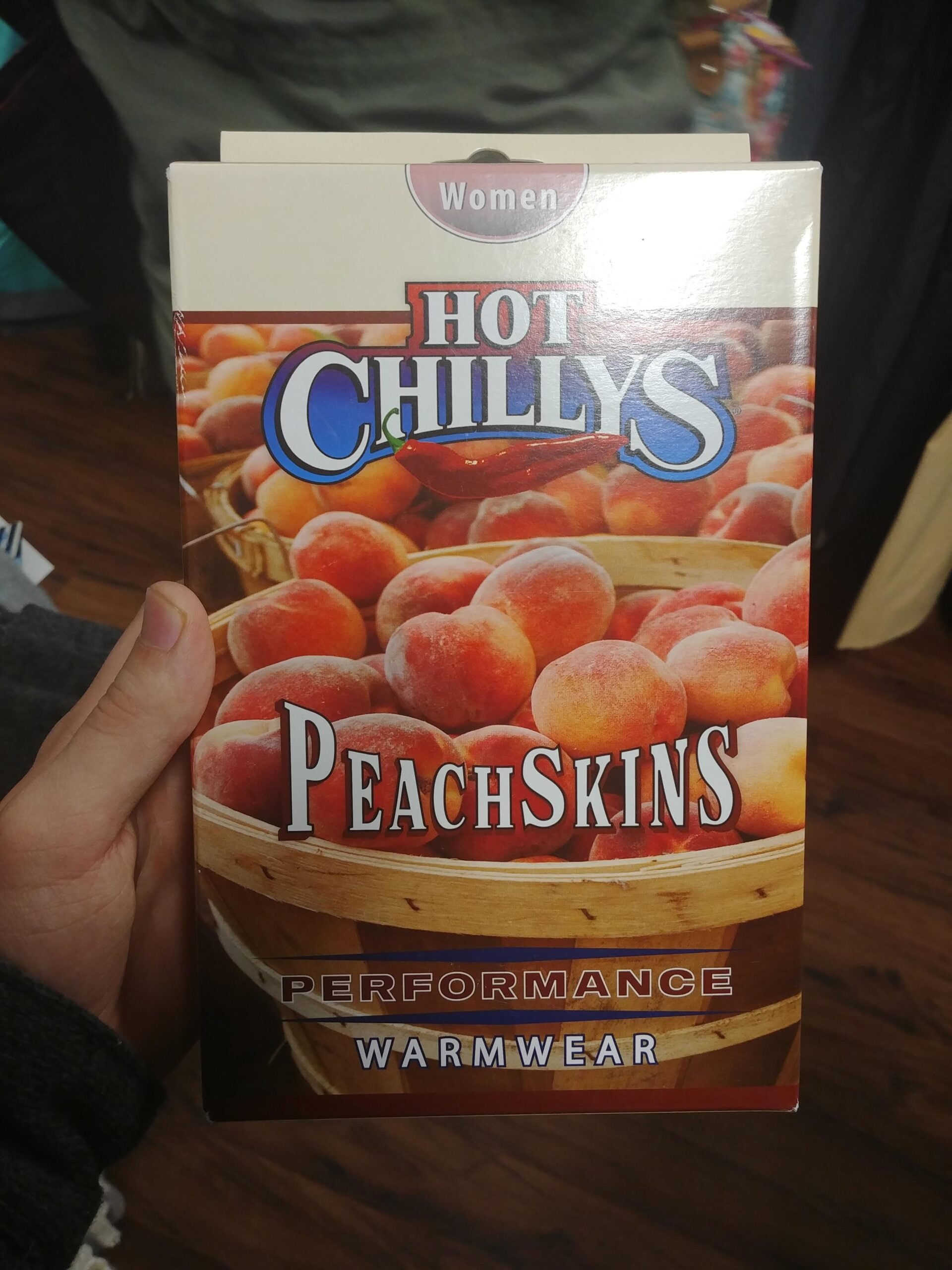 A hand holds a box labeled “Hot Chillys Peachskins Performance Warmwear” with an image of peaches in a basket and the word “Women” at the top.