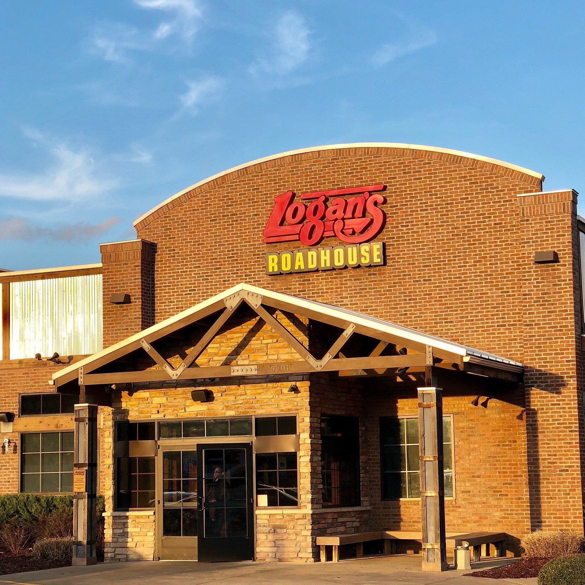 The exterior of a Logan’s Roadhouse restaurant, with a brick facade, large windows, and the Logan’s Roadhouse red and yellow sign above the entrance.
