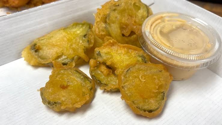 Five pieces of deep-fried pickle chips are served on a white tray next to a small plastic container of creamy dipping sauce.