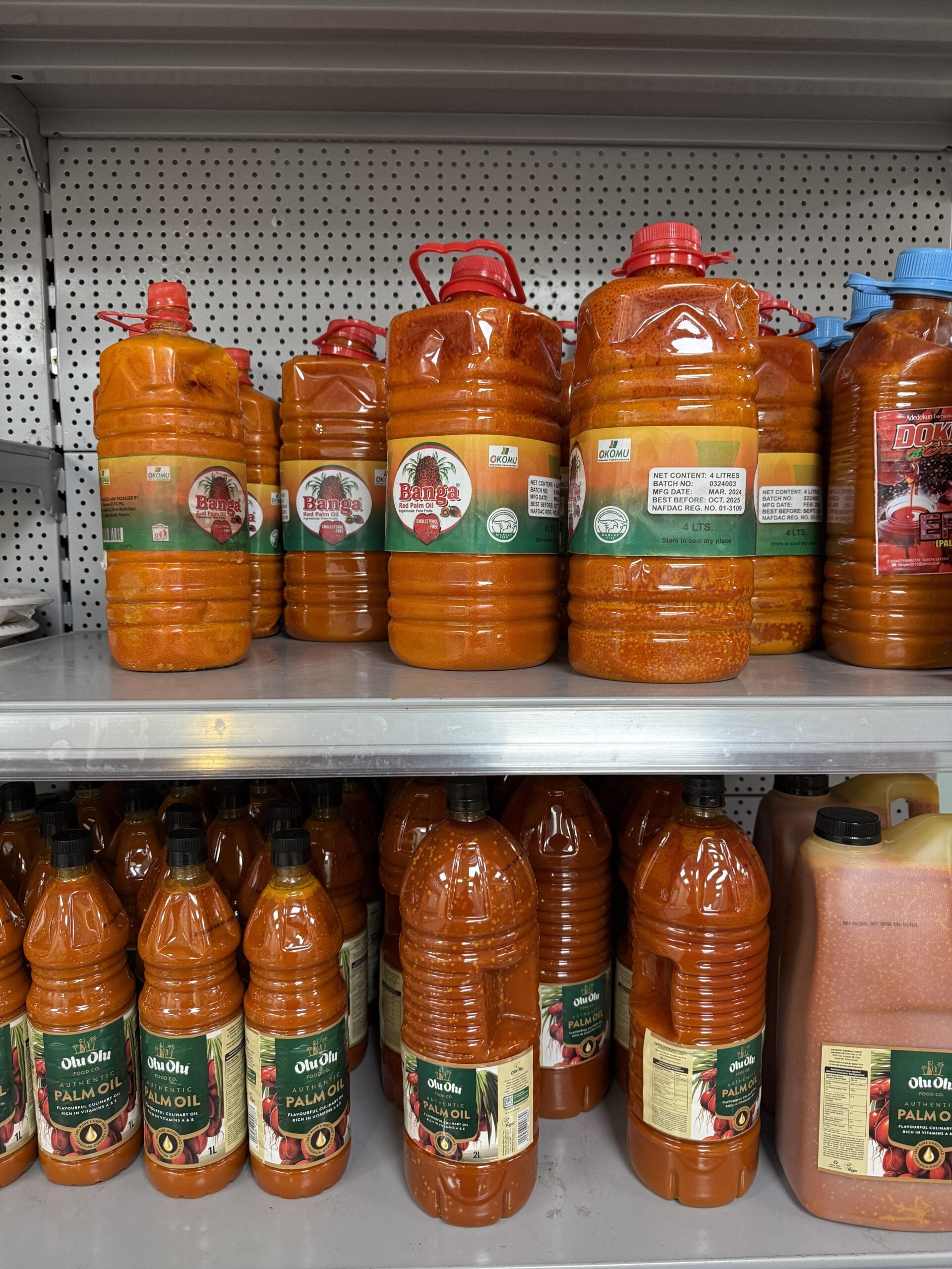Large plastic bottles of bright orange-red palm oil are displayed on metal shelves in a store. The bottles have red caps and green and white labels, with some bottles slightly stained with oil.