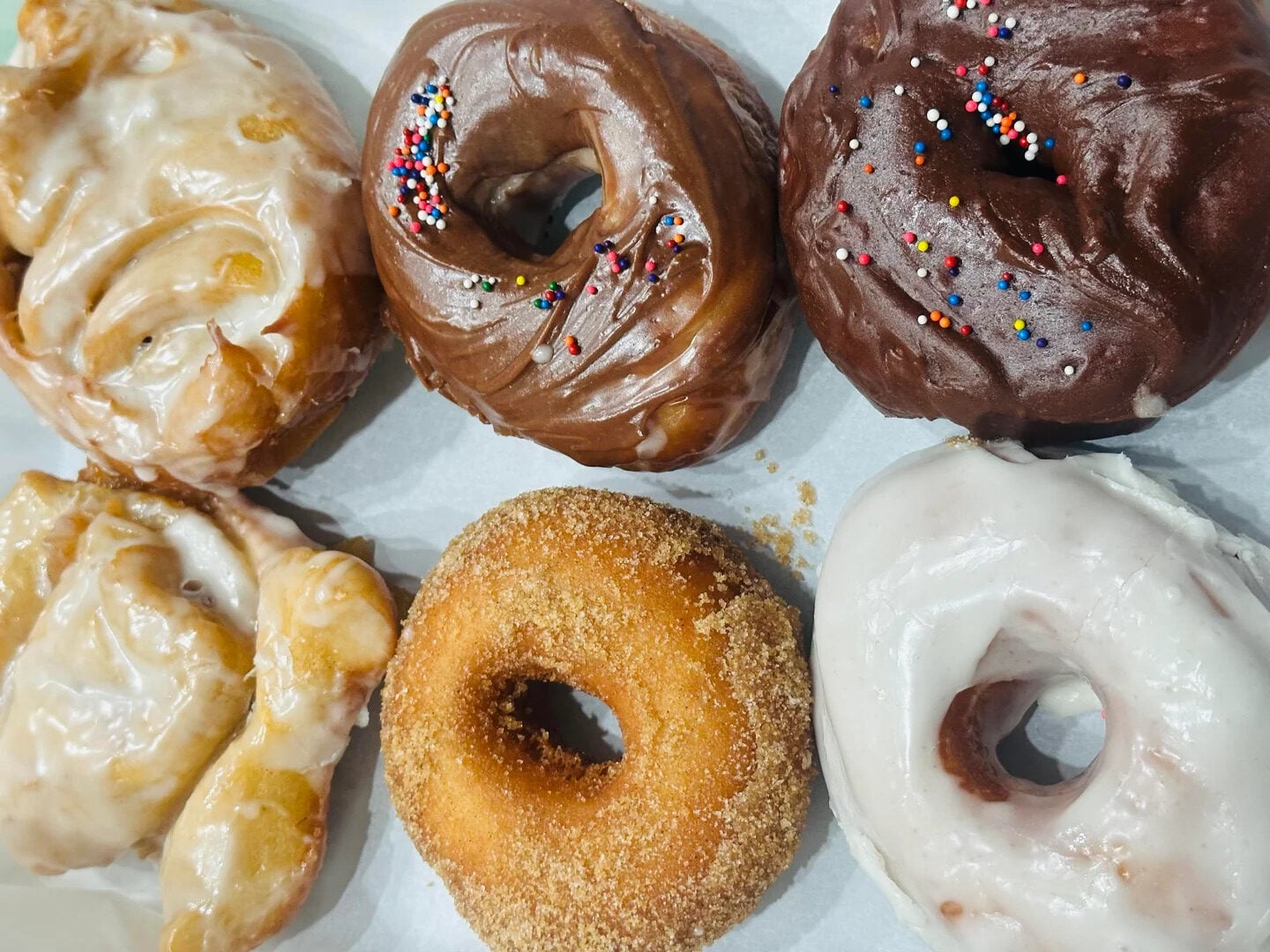 Six assorted donuts on a white surface: two glazed apple fritters, two chocolate frosted with sprinkles, one cinnamon sugar, and one classic glazed donut.