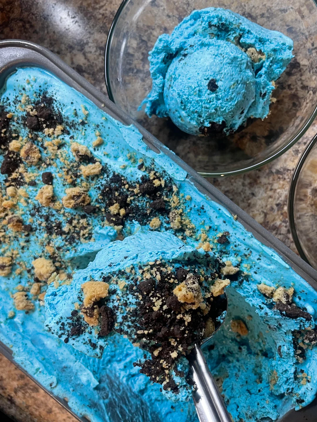 A close-up of bright blue ice cream with crushed cookies on top in a metal container, with a scoop served in a glass bowl nearby.