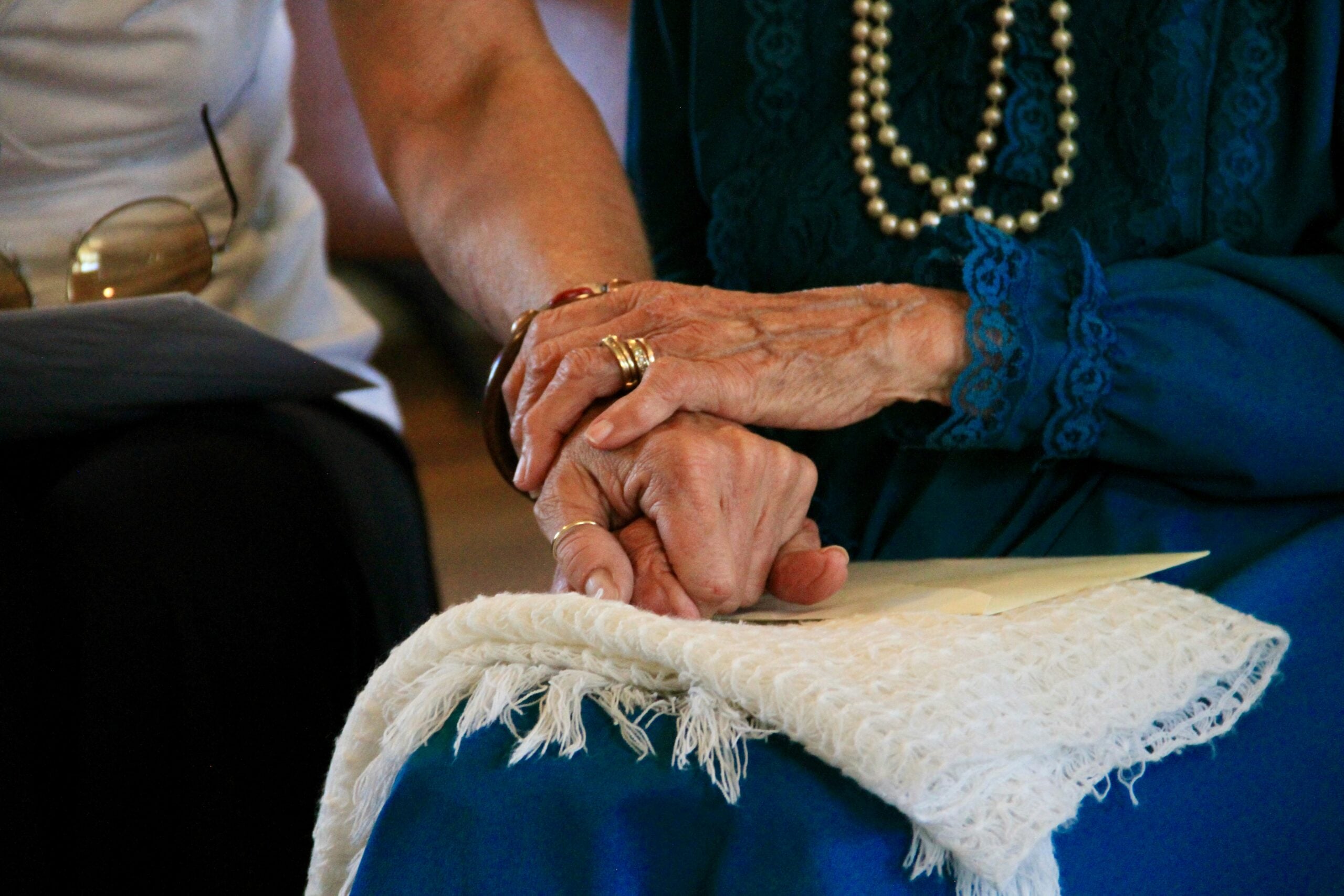 Two elderly women sit closely, one in blue with pearls, the other in white, gently holding hands—a tender moment that captures the essence of affection and support often celebrated in boomer facts. The focus is on their hands resting atop a fringed white cloth draped over their laps, emphasizing a lifetime of care and companionship.
