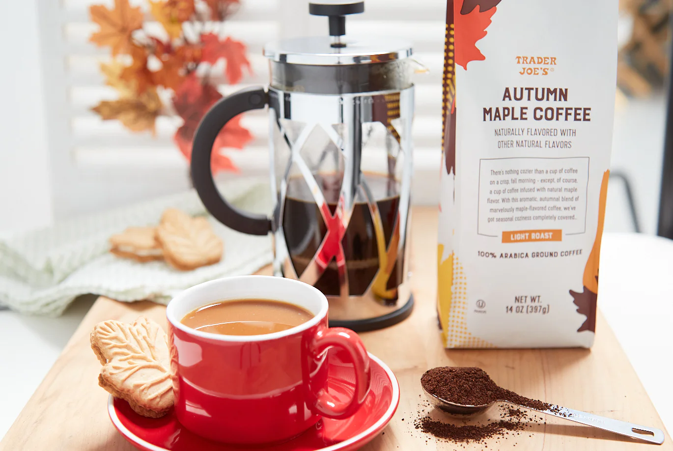 A red cup of coffee sits on a wooden tray with leaf-shaped cookies, coffee grounds, and a French press. Trader Joe’s Autumn Maple Coffee box is nearby. Autumn leaves are in the background.
