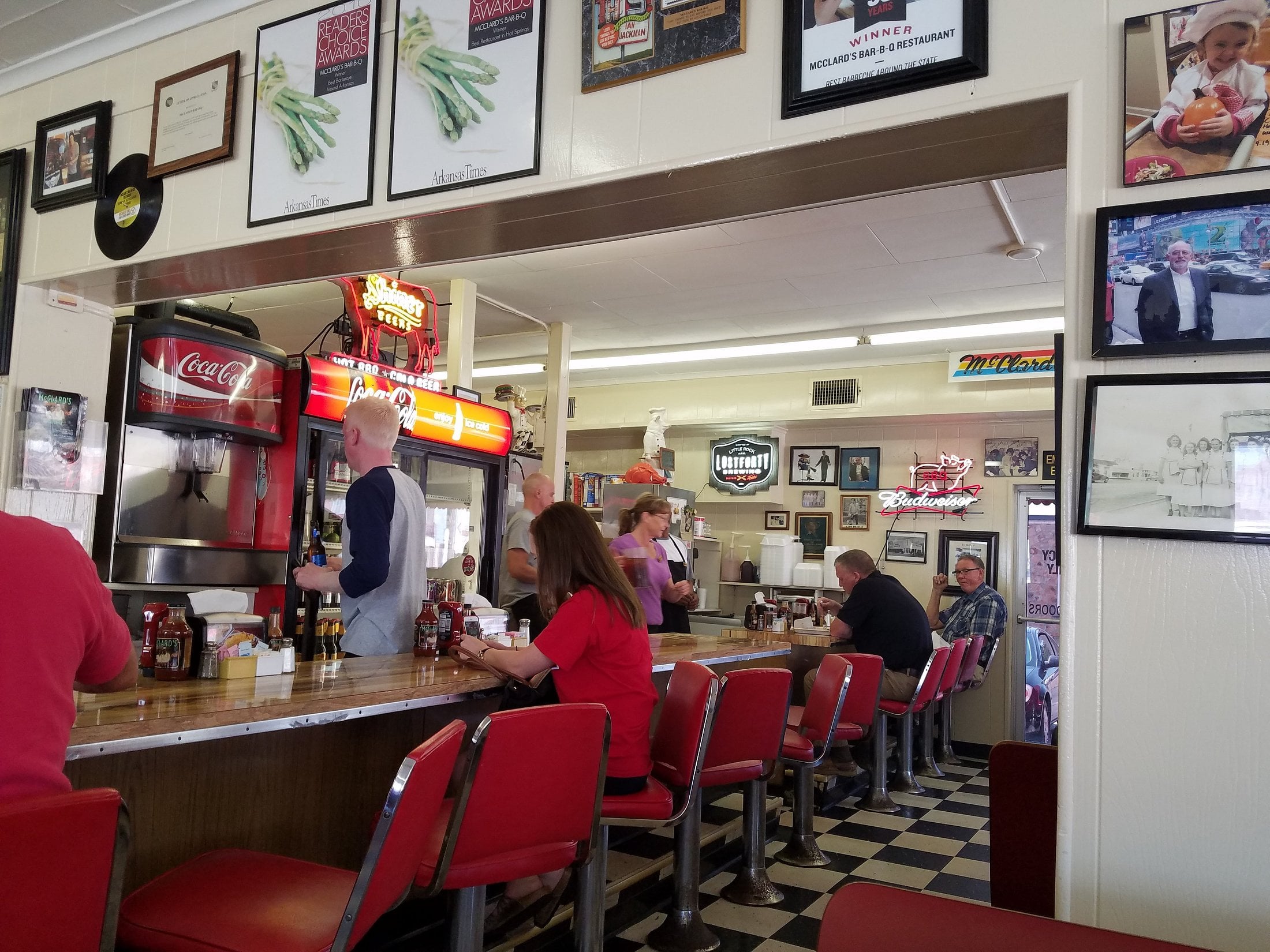 Classic diner scene with red stools, people sitting at the counter, and a server taking orders. Walls are decorated with photos and signs. A Coca-Cola soda fountain and checkered floor are visible.