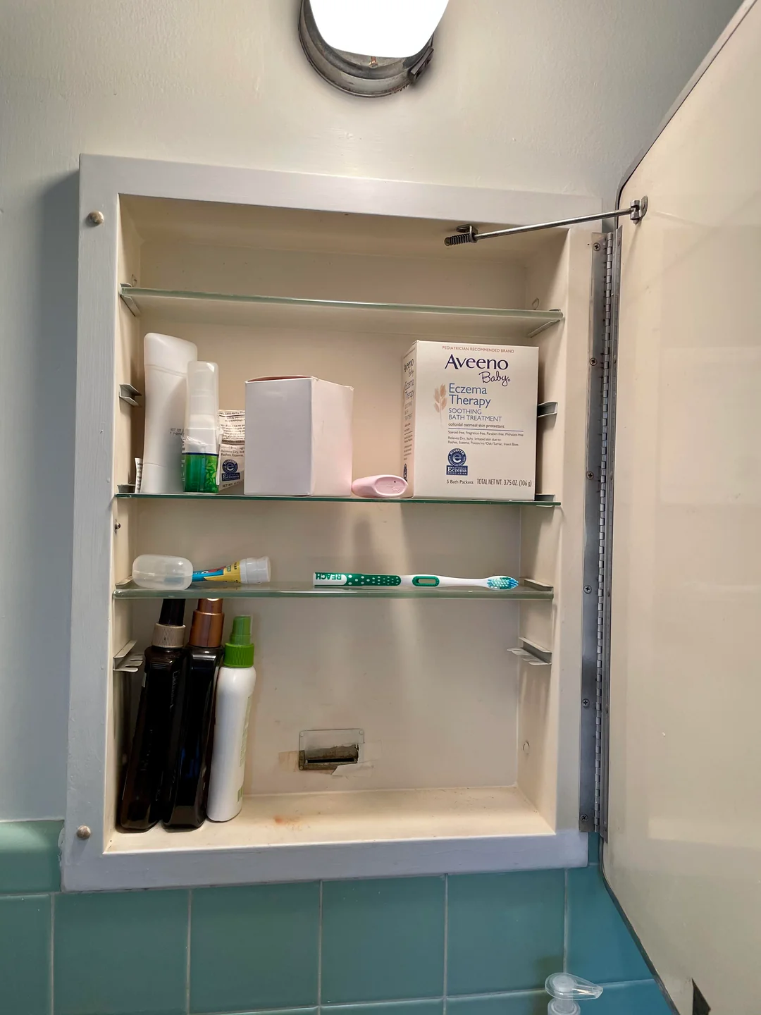 An open bathroom medicine cabinet with shelves holding a box of Aveeno cream, bottles, a toothbrush, toothpaste, a tube, and other toiletries. The cabinet is white with a mirrored door.