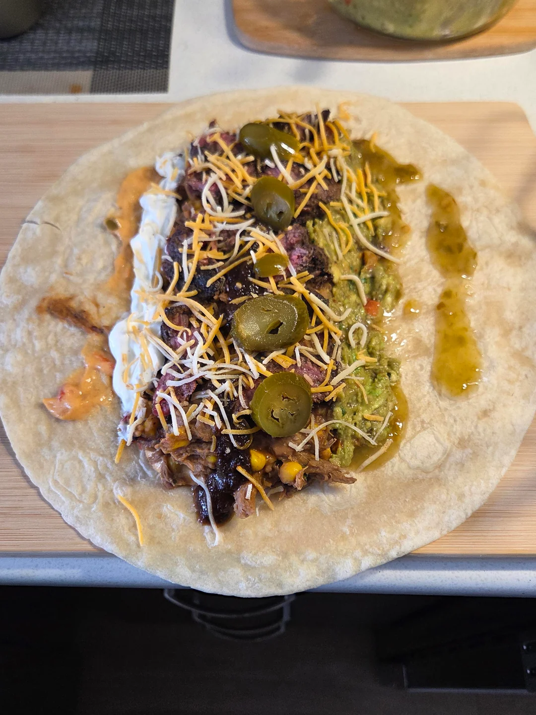 A tortilla topped with shredded meat, black beans, corn, shredded cheese, guacamole, jalapeños, sour cream, salsa, and green sauce, ready to be wrapped into a burrito, on a wooden surface.