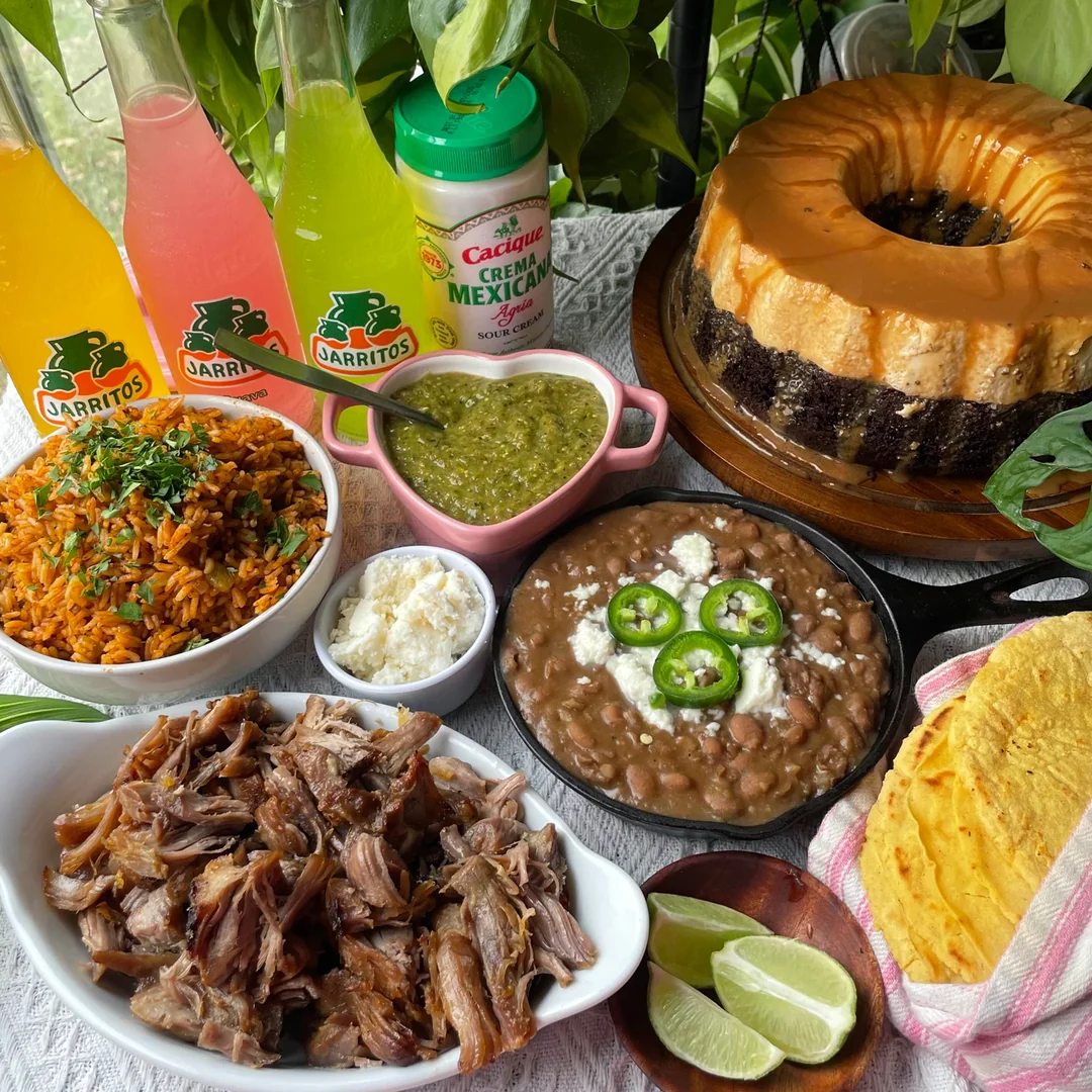 A table with Mexican food: carnitas, rice, beans, tortillas, green salsa, lime wedges, crema, three bottles of Jarritos soda, and a marble bundt cake, surrounded by green leaves.