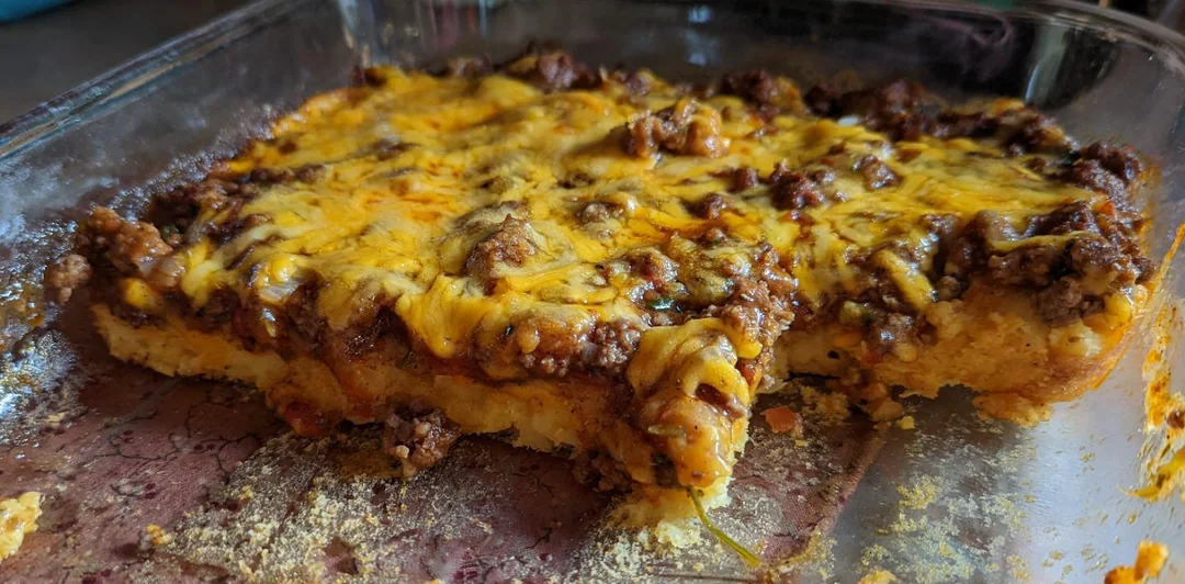 A homemade casserole in a glass baking dish, topped with melted cheddar cheese and ground beef, with a large portion removed, revealing a thick, soft crust underneath.