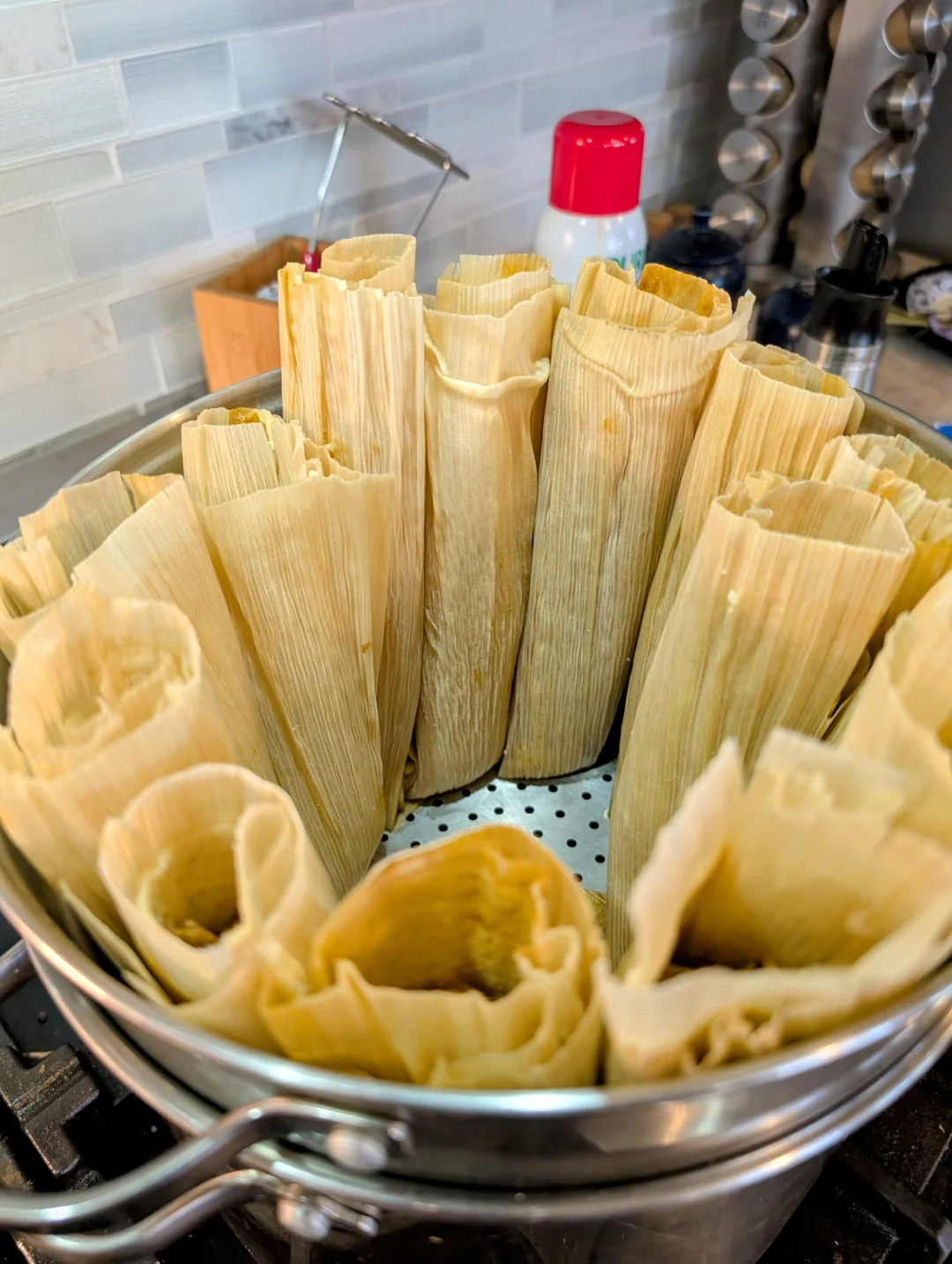 A pot of tamales wrapped in corn husks stands upright inside a steamer basket on a stovetop, ready to be cooked. The kitchen backsplash and various utensils are visible in the background.