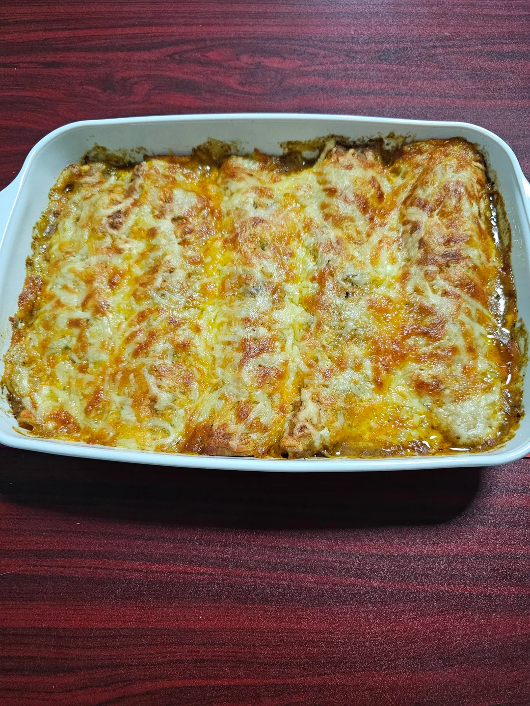 A baked dish of cheesy lasagna in a white rectangular casserole pan, with melted golden-brown cheese and tomato sauce visible on top, on a red wooden surface.