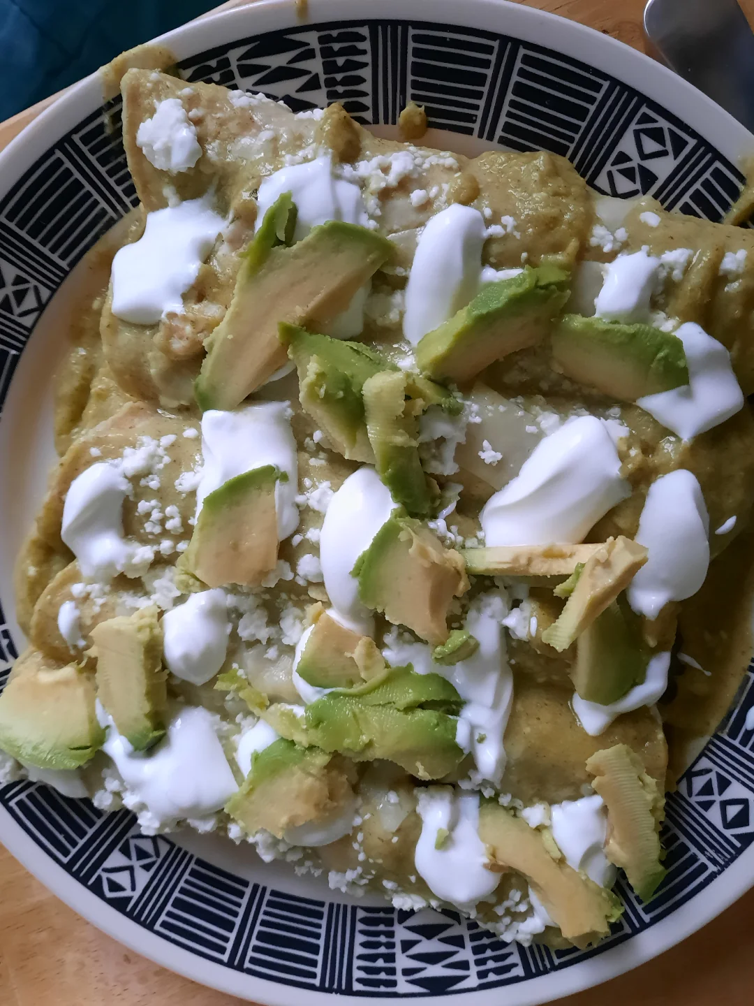 A plate of enchiladas covered in green sauce, topped with sliced avocado, sour cream, and crumbled cheese, served on a patterned plate.