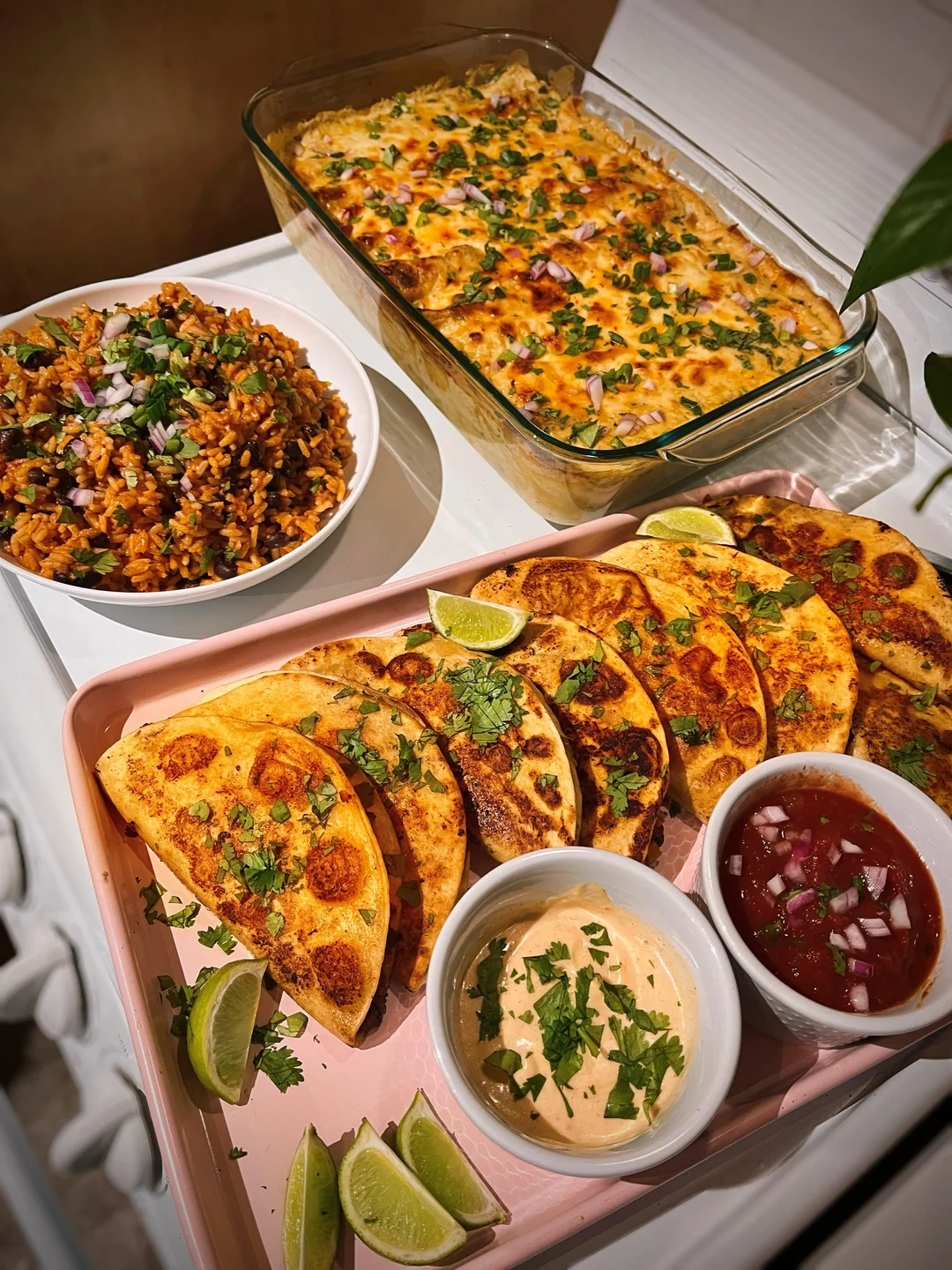 A tray of folded tacos with dips and lime wedges, a dish of cheesy casserole, and a bowl of seasoned rice topped with onions. Food is garnished with fresh herbs, arranged on a stove and a pink tray.