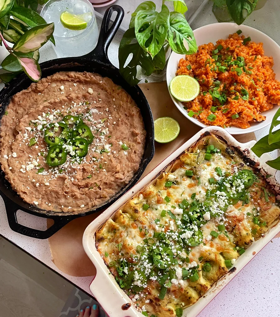 A skillet of refried beans topped with jalapeños, a dish of cheesy enchiladas, and a bowl of Mexican rice garnished with cilantro and lime, set on a countertop with potted plants nearby.