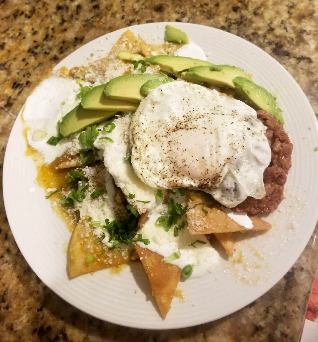 A plate of chilaquiles topped with sliced avocado, a fried egg, refried beans, crema, cheese, and cilantro is on a granite countertop.
