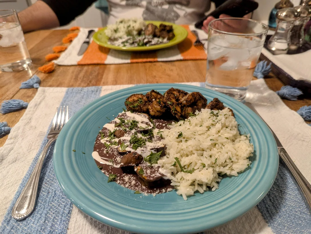 A plate with rice, grilled chicken, and black beans topped with cream and herbs sits on a set table. A fork is on the plate, and a glass of water is beside it. Another plate is visible across the table. Homemade Mexican food.