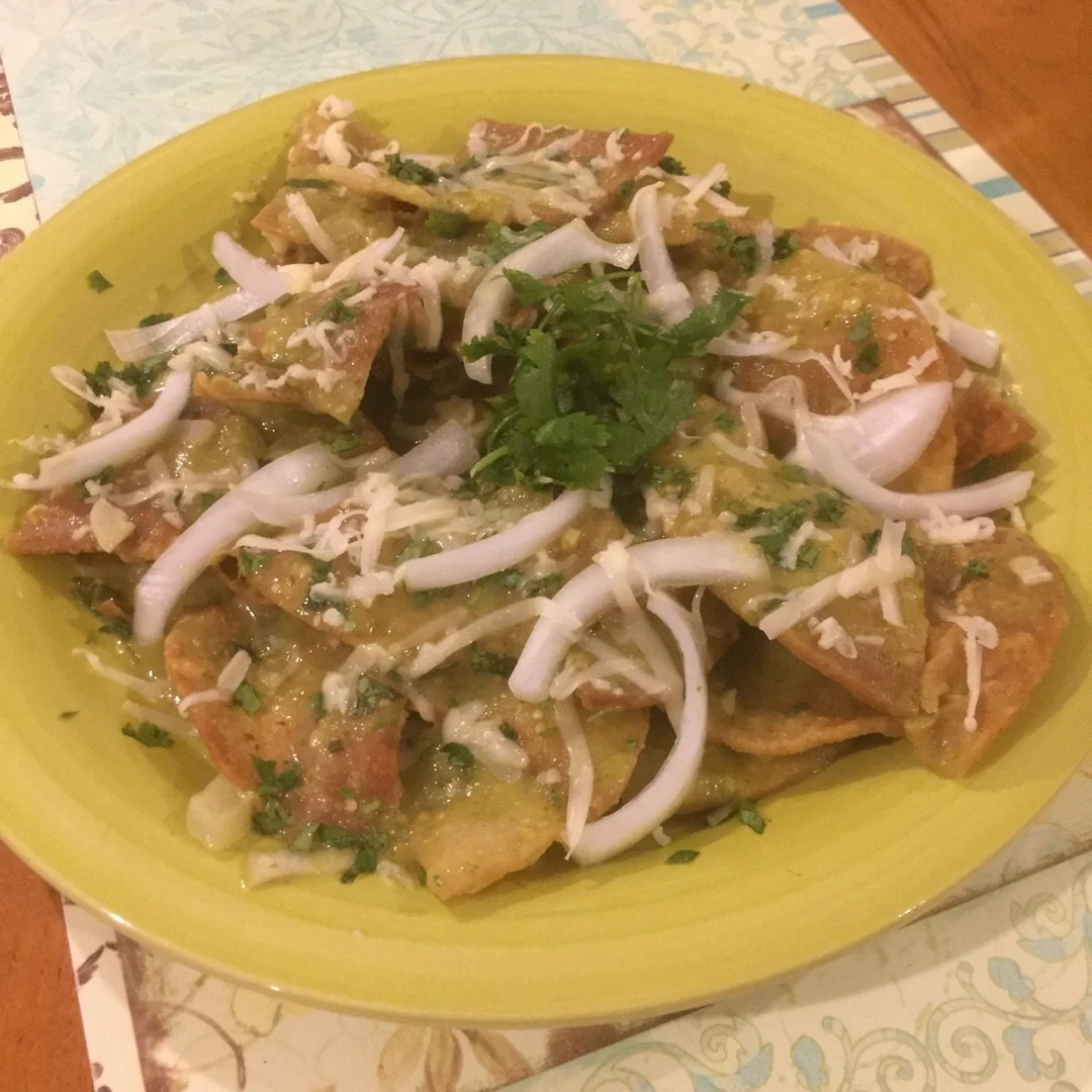 A yellow plate with chilaquiles topped with green salsa, sliced onions, shredded cheese, and chopped cilantro, on a table with placemats.