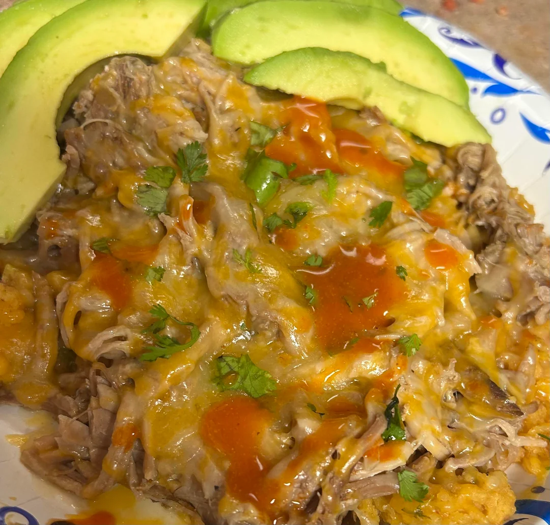 Shredded meat topped with melted cheese, hot sauce, and chopped cilantro, served with sliced avocado on a plate.