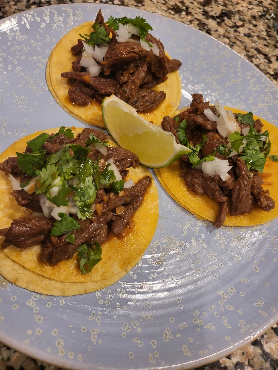 Three street tacos with chopped beef, onions, and cilantro are served on a blue plate with a wedge of lime. The tacos are on yellow corn tortillas, and the plate sits on a granite countertop.
