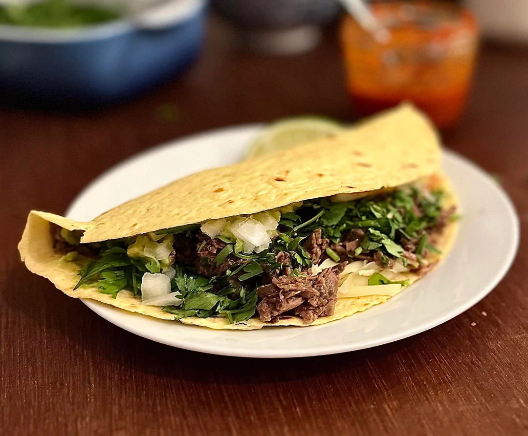 A large taco filled with shredded meat, chopped onions, and fresh cilantro sits on a white plate, with a lime wedge and blurred salsa jar in the background.