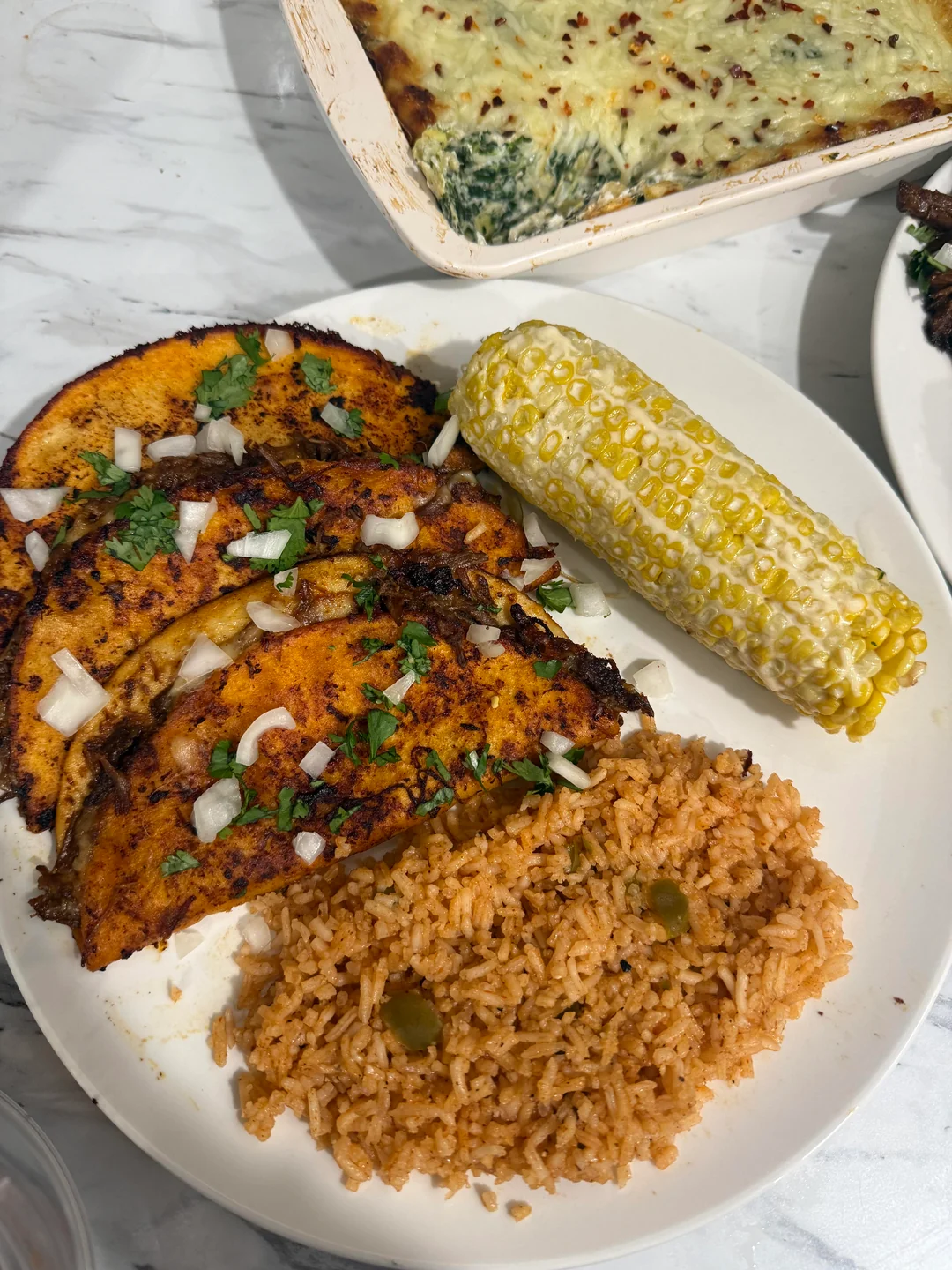 A plate with three birria tacos topped with onions and cilantro, a serving of Mexican rice with peas, and a cob of corn. In the background is a spinach lasagna dish.