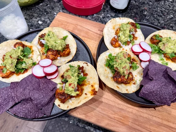 Two plates with tacos topped with guacamole, cilantro, and grilled meat, each served with sliced radishes and blue corn tortilla chips, on a dark countertop with a wooden cutting board underneath.