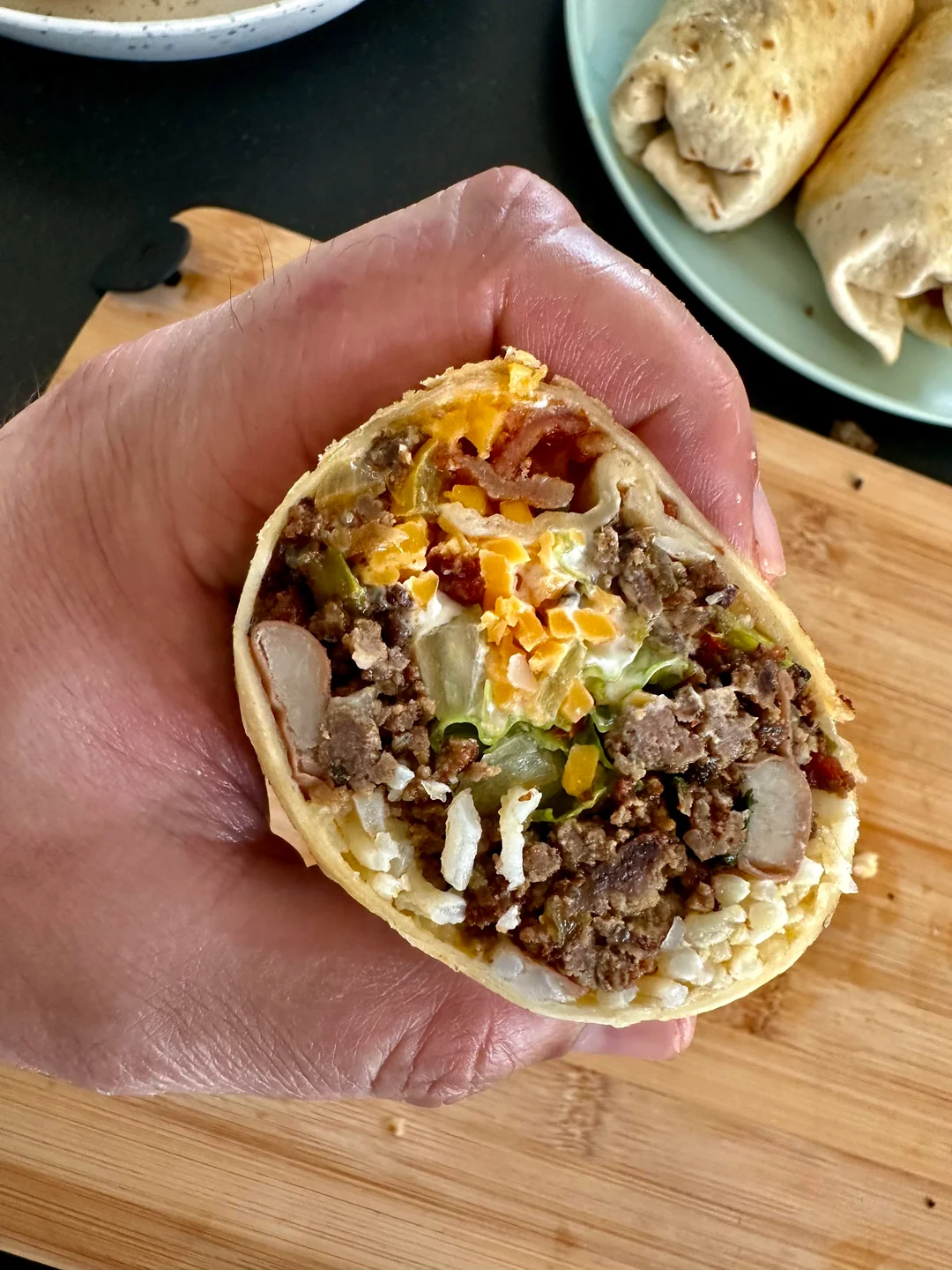 A hand holds a burrito cut in half, showing ground beef, beans, rice, lettuce, and shredded cheese inside. Other burritos are visible on a plate and a wooden board in the background.