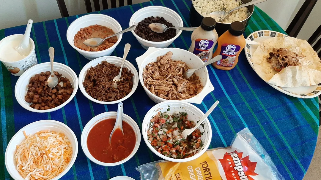 A table with various taco ingredients: shredded chicken, ground beef, beans, salsa, pico de gallo, shredded cheese, rice, sauces, sour cream, and tortillas on a blue plaid tablecloth.