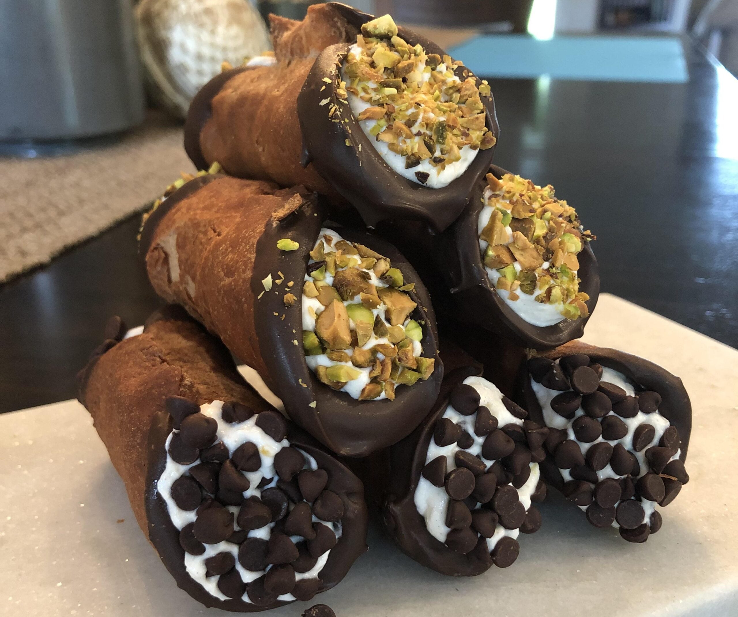 Six homemade cannolis stacked in a pyramid, with ends dipped in chocolate and coated with chopped pistachios or mini chocolate chips, on a marble surface.