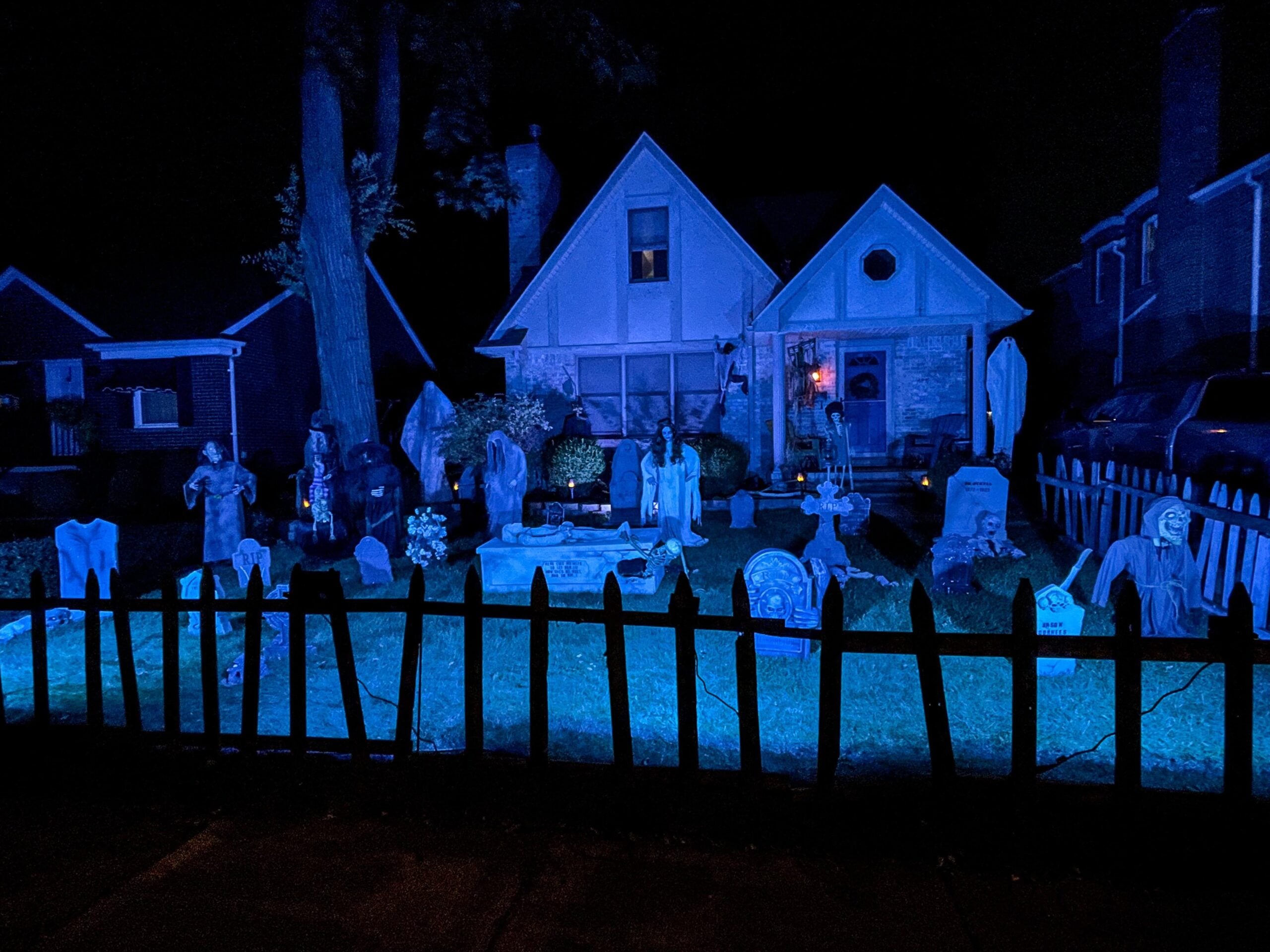 A house and yard are decorated with blue lighting, tombstones, skeletons, and eerie props, showcasing Halloween yard decorations that create a spooky graveyard scene at night.