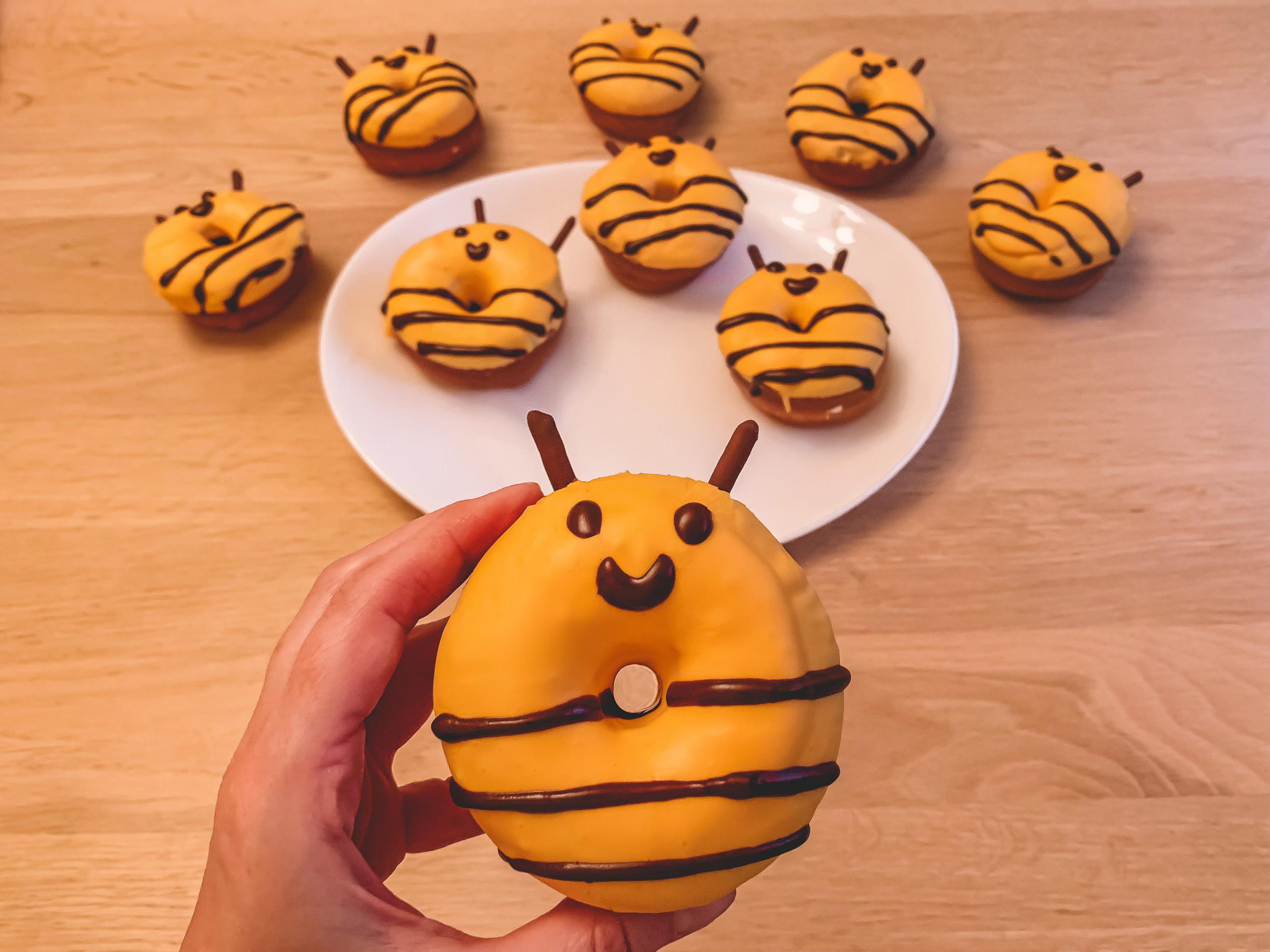 A hand holds a bee-shaped donut with yellow icing, chocolate stripes, and candy eyes. More bee donuts are arranged on a white plate and the wooden table in the background.