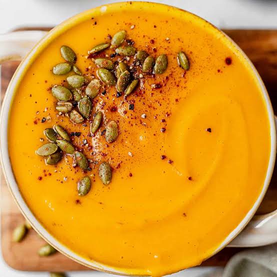 A bowl of creamy orange soup topped with roasted pumpkin seeds, cracked pepper, and spices, placed on a wooden surface. Fall comfort foods under $10.