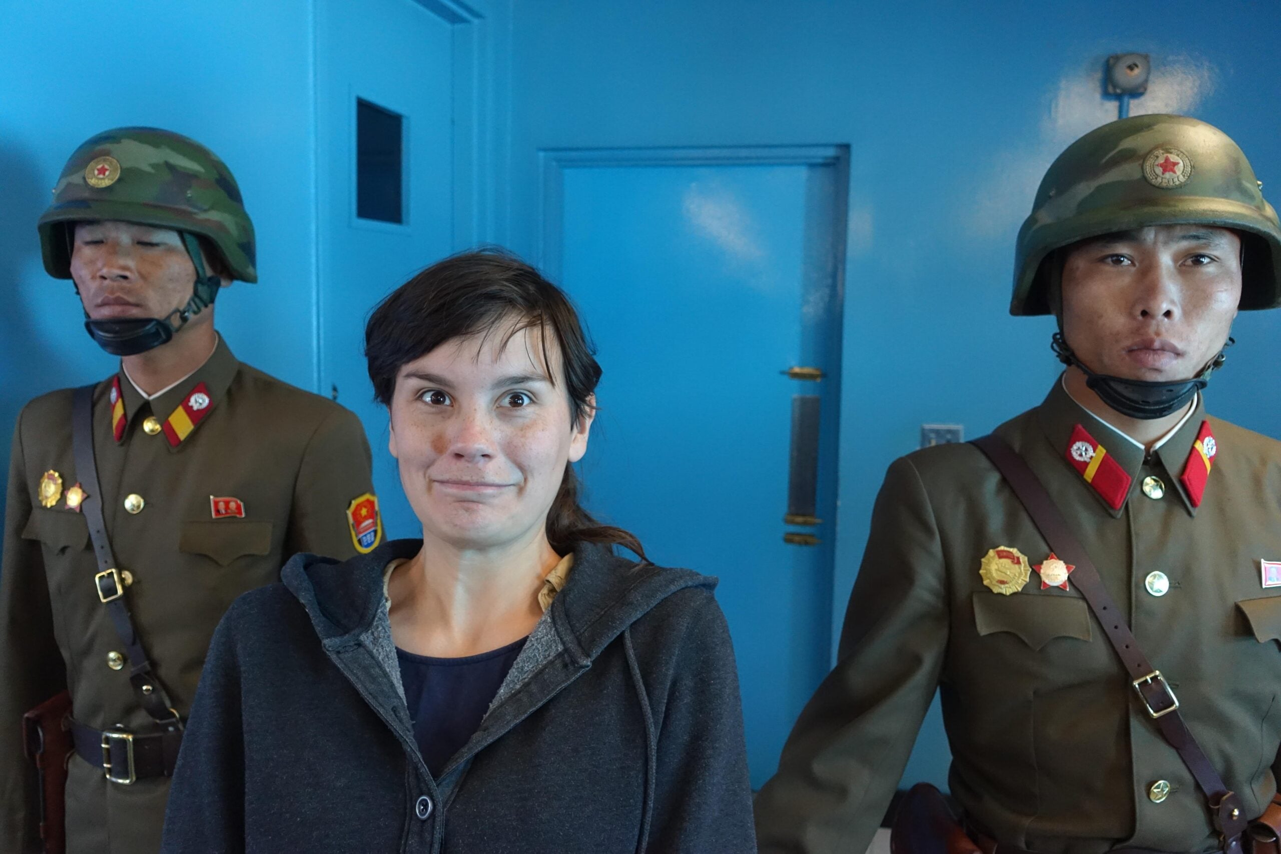 A woman stands between two North Korean soldiers in uniform and helmets, inside a blue-walled room. The woman is making a surprised face while the soldiers remain expressionless.